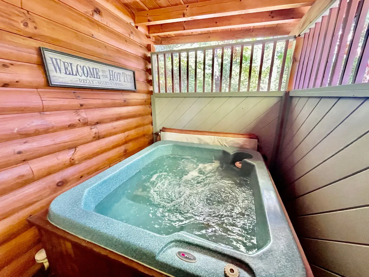 A tranquil interior view of the private, fully enclosed hot tub area, framed by rustic wooden walls and a friendly welcome sign. The warm lighting and intimate setting make it perfect for unwinding.