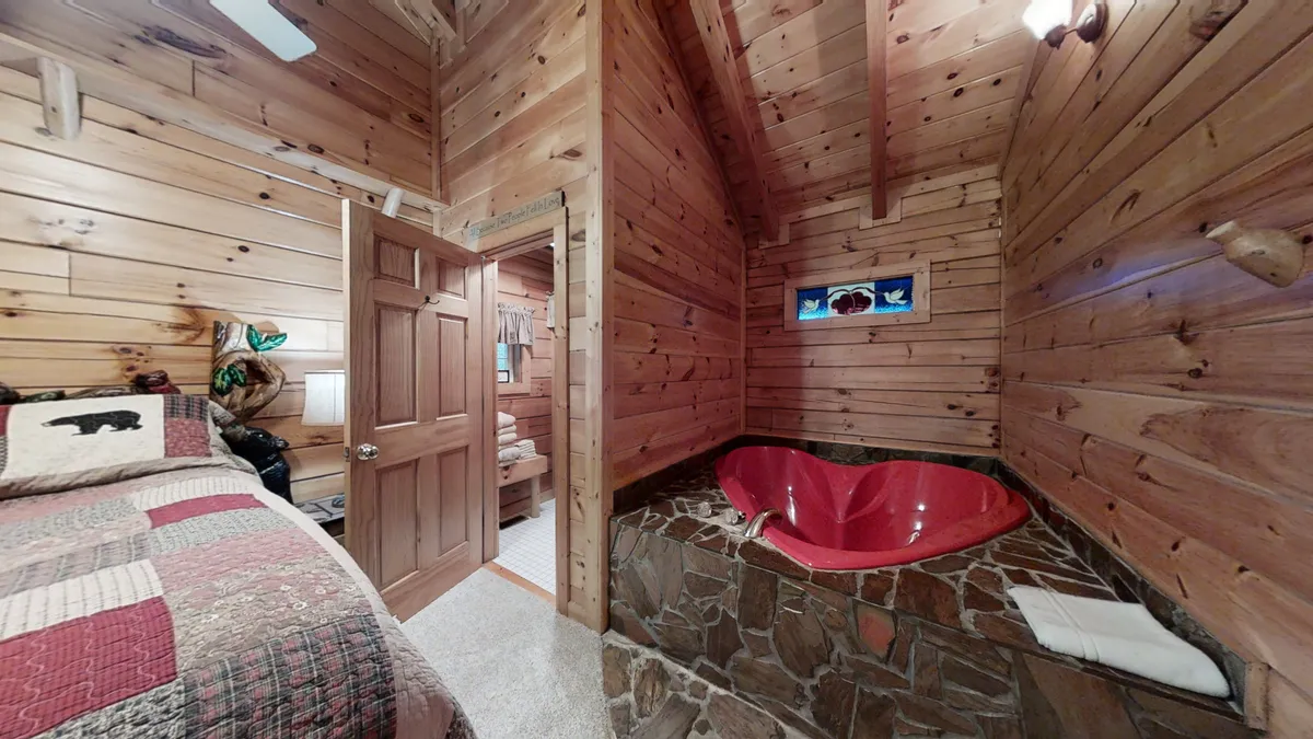 A heart-shaped red jacuzzi tub is the showstopper in this image, set against textured wooden walls. The bed with plaid bedding is visible nearby, and nature-themed accents complete the tranquil, private atmosphere. The tub's proximity to the bedroom emphasizes both luxury and intimacy.