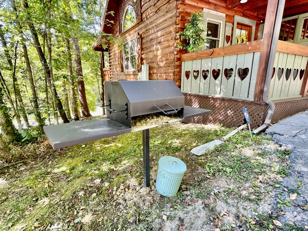 An exterior shot capturing the wooden cabin with a charcoal grill ready for outdoor cooking. Heart-shaped cutouts decorate the porch railing, while lush green foliage completes the cozy, mountain retreat feel.