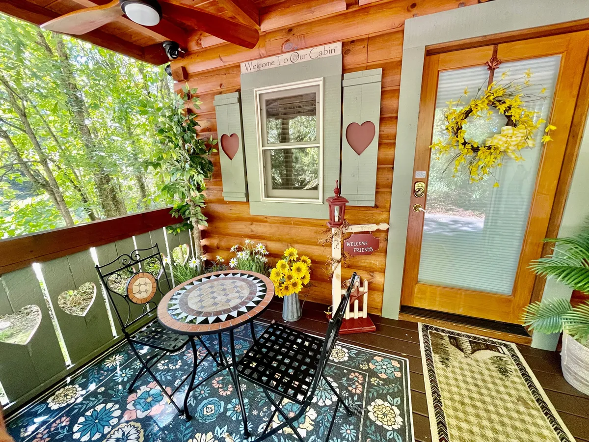 A cozy exterior view of the front porch, featuring a mosaic-topped table with black chairs, a welcoming sign, a vase of sunflowers, and a decorative wreath. The space is surrounded by greenery, creating a relaxing spot to enjoy morning coffee or evening conversations.