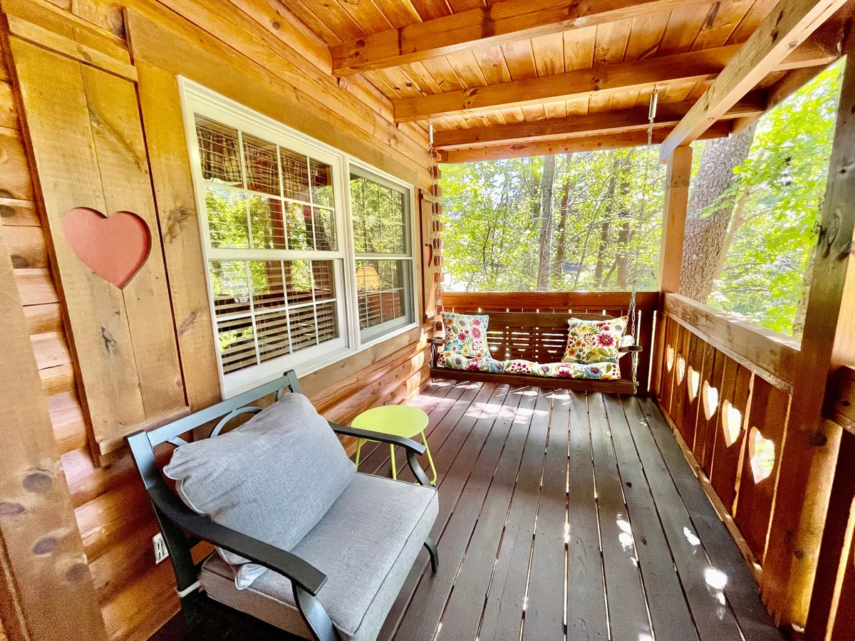 A cozy swing bench adorned with colorful cushions sits beside a gray armchair in a bright corner with large windows and a wooden wall with a heart cutout. The setting merges indoor comfort with clear views of the outdoors, inviting guests to relax while remaining connected to nature.