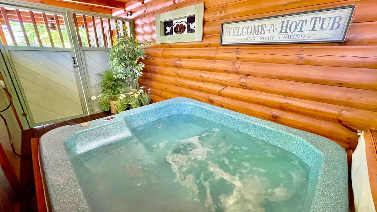 The image showcases the inviting interior of a cozy cabin, with a bubbling hot tub as the centerpiece, surrounded by lush green plants and a cheerful welcome sign. Soft natural light filters in, setting a tranquil, welcoming mood ideal for relaxation and connection.