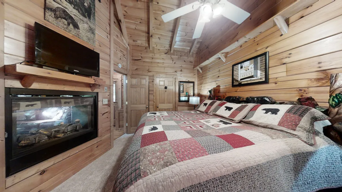 This image features a king-sized bed with a cozy quilt, wooden walls, a ceiling fan above, and a private gas fireplace glowing warmly at the foot of the bed. The inviting space exudes comfort and rustic charm, ideal for couples seeking romance and relaxation, with a TV visible for added entertainment.