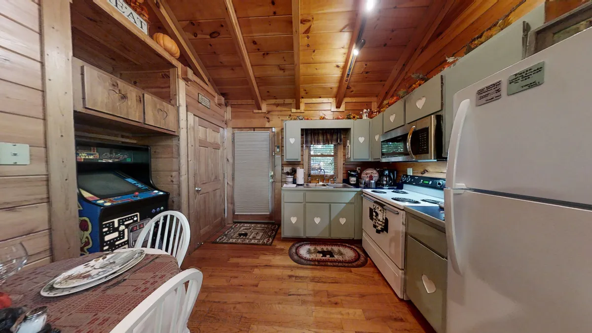 This image captures a warm, nostalgic kitchen and dining area. The wooden cabinetry and white appliances create a homey atmosphere, while the dining table is ready for casual meals. Most notably, a retro arcade game is visible in the background, hinting at playful gatherings and classic family fun.
