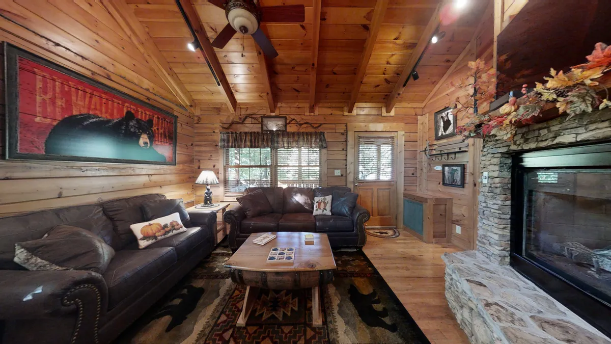 This image showcases a warm and inviting living room featuring plush leather sofas, a beautiful stacked-stone fireplace, a wooden ceiling, and a coffee table with magazines. The decorative bear painting above the fireplace adds a charming, rustic touch, while the overall ambiance feels perfect for relaxing evenings by the fire or cozy gatherings.