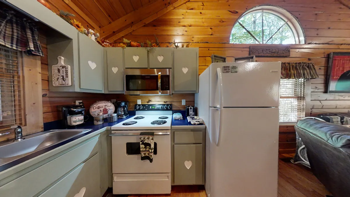 A spacious kitchen area equipped with modern appliances and charming heart-shaped cabinet designs. The wooden ceiling and a large circular window create a cozy, sunlit environment ideal for both cooking and gathering with family or friends around the dining table.
