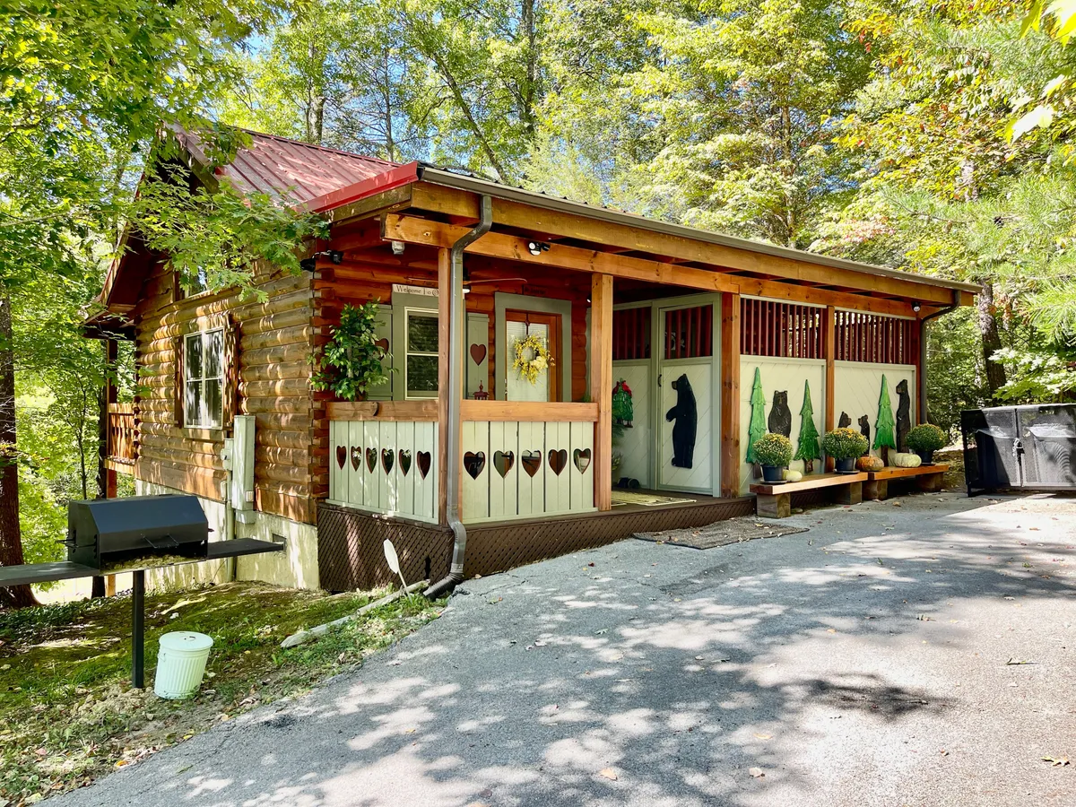 A charming wooden porch adorned with bear motifs and heart-shaped cutouts, surrounded by lush greenery. The porch features a cozy sitting area perfect for relaxing, with open views that connect the cabin to the outdoors.