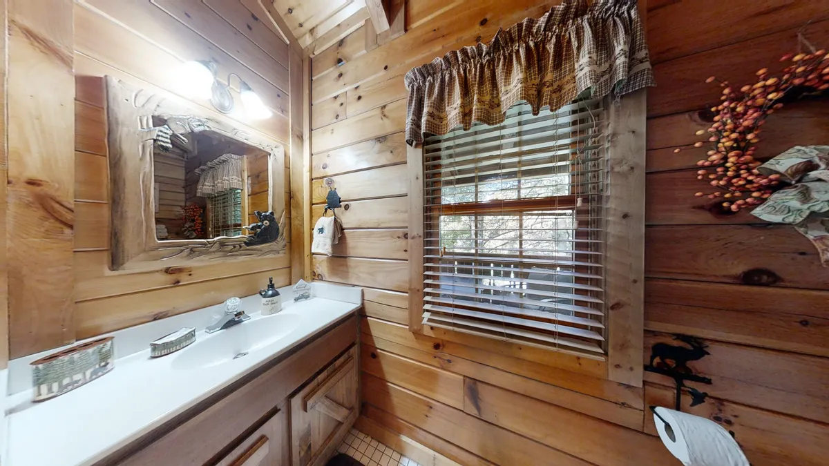 This image highlights the ensuite bath area, featuring a rustic wooden mirror, a well-lit sink area, and a window with blinds. Soft light streams in, reflecting off the wooden interiors to create a serene retreat for personal care.