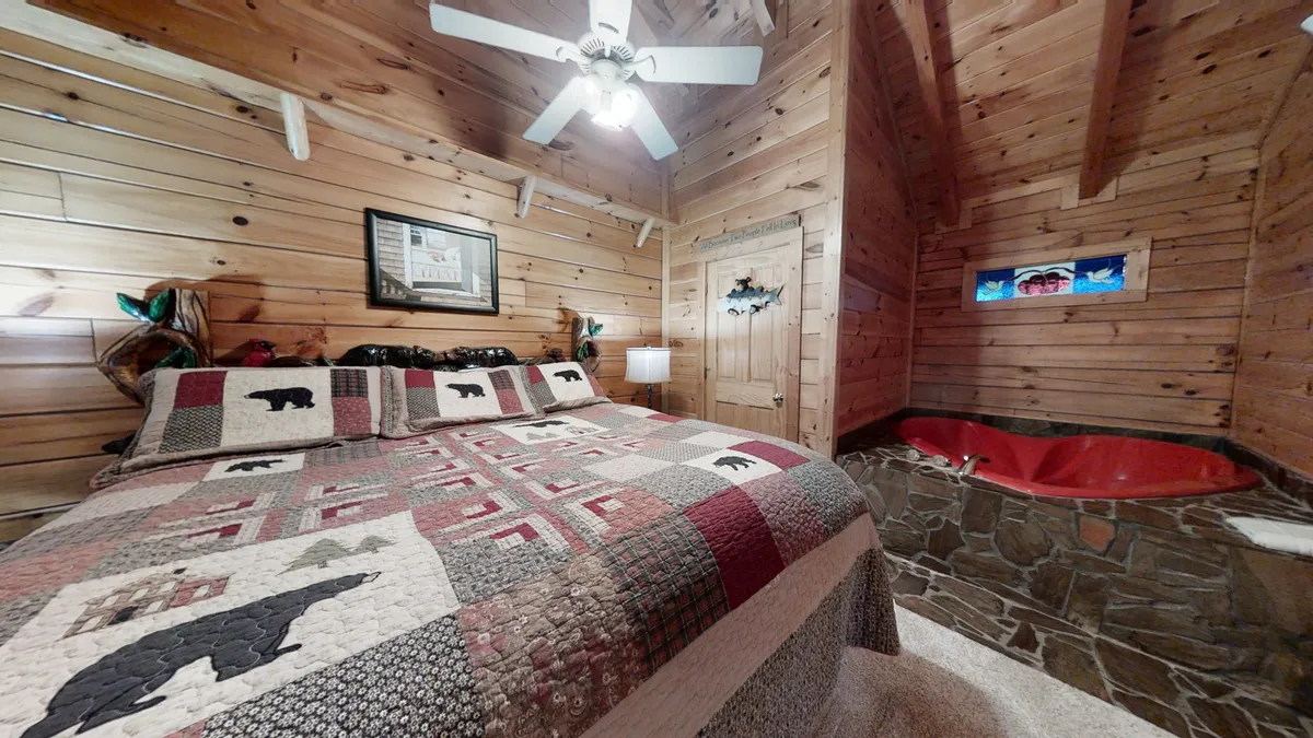 A cozy interior with a quilted bedspread featuring bear motifs, a unique red heart-shaped hot tub, and wooden walls. The space is intimate and inviting, designed for relaxing soaks and snuggling up together.